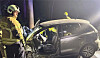 Feuerwehrleute in Schutzkleidung begutachten ein schwer beschädigtes Auto am dunklen Straßenrand.