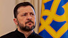 Volodymyr Zelensky stands beside a Ukrainian trident emblem in blue and yellow.