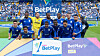 Jugadores de Millonarios posan en el campo junto a un panel de BetPlay.