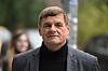 An unidentified man in a grey jacket and black jumper stands outdoors, with blurred people and trees behind him.